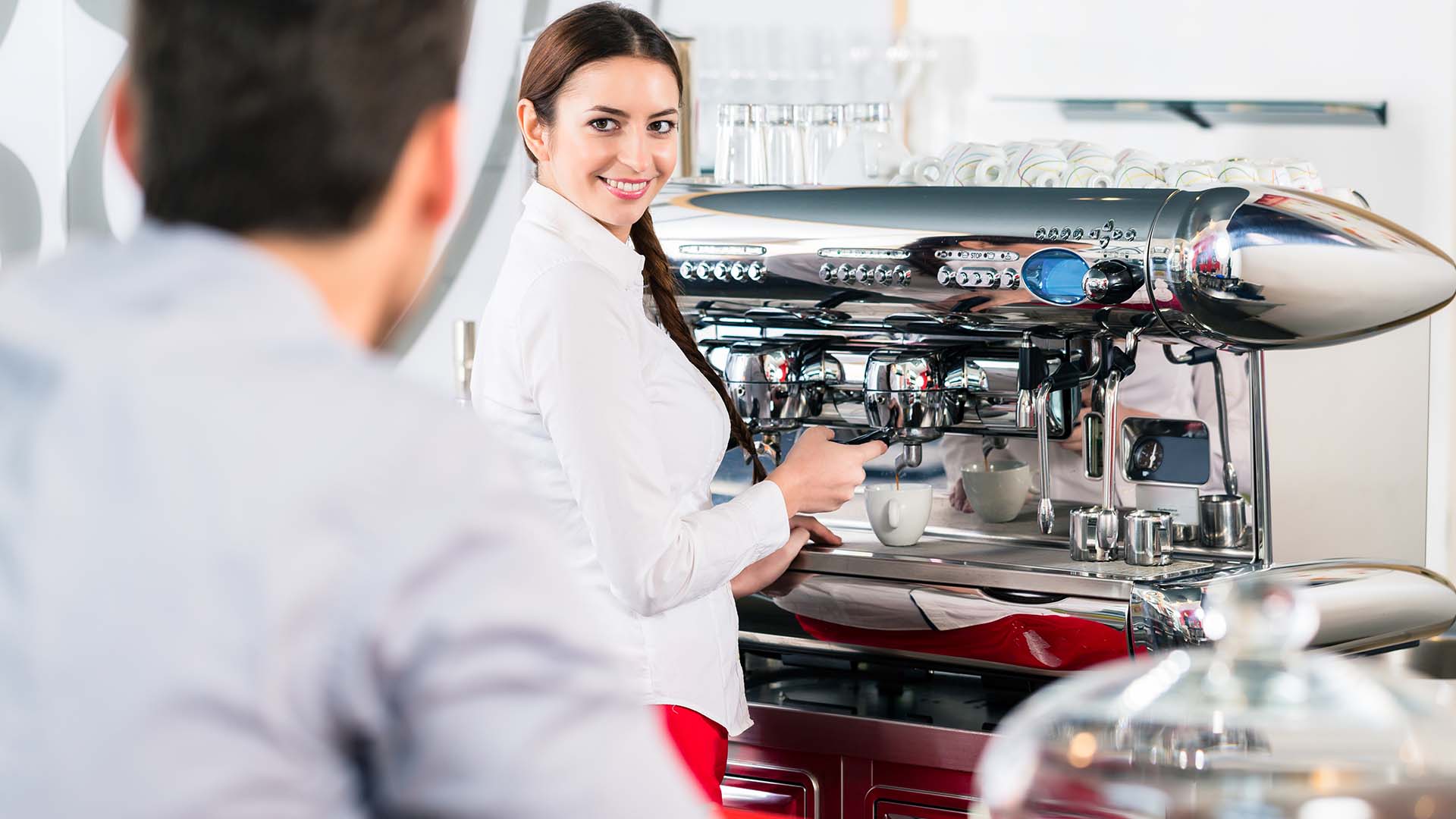 Brunette woman and man conversing and smiling at each other while woman is preparing a coffee in front of a coffee machine.